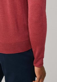 Červený svetr s dlouhým rukávem a texturovanými nášivkami na loktech, nošený přes tmavě modré kalhoty, zobrazuje záda a ruku osoby.