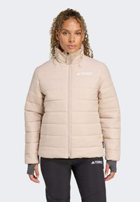 Kvinde med krøllet hår iført en lys beige pufferjakke med højt krave, står mod en neutral baggrund, hænderne hvilende langs hendes sider.