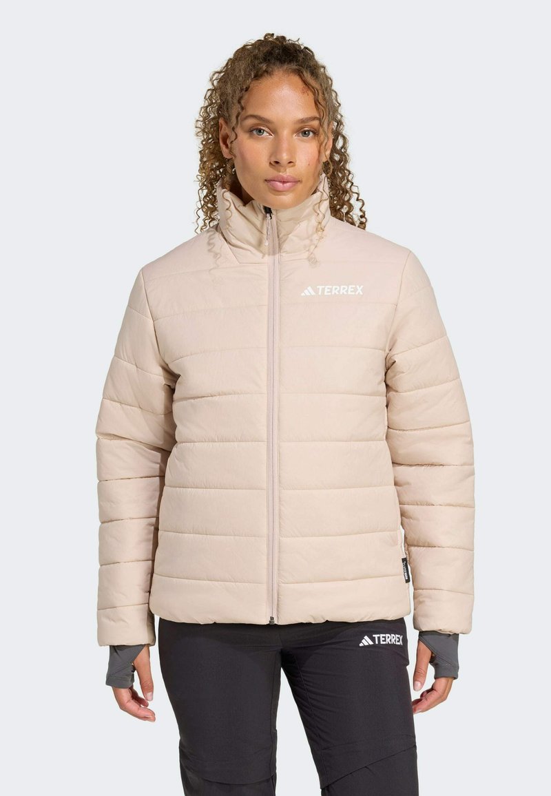 Kvinde med krøllet hår iført en lys beige pufferjakke med højt krave, står mod en neutral baggrund, hænderne hvilende langs hendes sider.