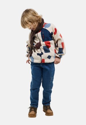 Enfant portant une veste en polaire crème avec des formes abstraites colorées, un jean cargo bleu, une écharpe marron et des bottes beige, regardant vers le bas.