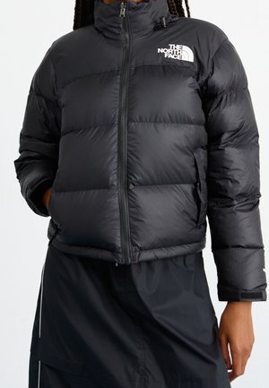 Vrouw draagt een zwarte pufferjas met het The North Face-logo op de borst en zwarte broek, handen in de jaszakken.