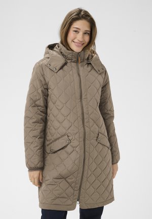 Ung kvinde smilende iført en lang quiltet beige frakke med hætte og forlommer, stående mod en ensfarvet hvid baggrund.