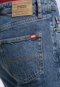 Jeans de mezclilla azul con un parche de cuero con la marca "Big Star", costuras amarillas en contraste y una pequeña etiqueta roja en el bolsillo trasero.