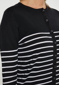 Cardigan a righe bianche e nere con scollo rotondo, caratterizzato da bottoni neri lungo la parte anteriore e una morbida texture a maglia.