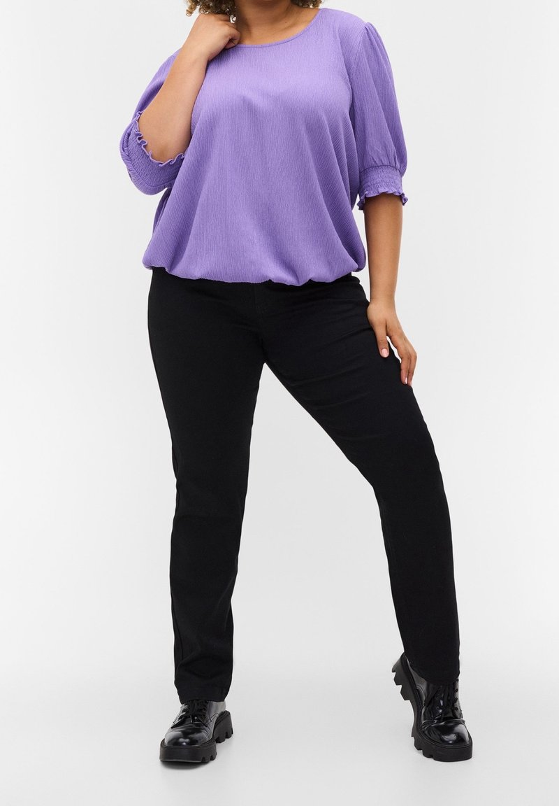 Femme portant un chemisier violet ample, un pantalon noir slim et des chaussures noires épaisses à lacets, debout avec une main près du cou et l'autre sur la cuisse.