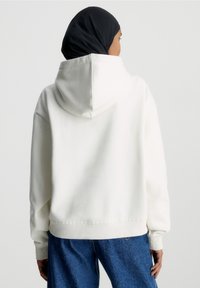 Woman wearing a black headscarf, white hooded sweatshirt, and blue jeans facing away against a plain light background.