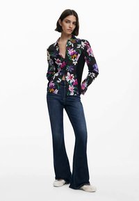 Zwart shirt met bloemenpatroon en lange mouwen, gecombineerd met uitlopende donkere denim jeans. Model staat tegen een witte achtergrond.