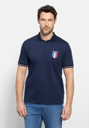 Man draagt een marineblauw polo met FRA-wapenpatch en gestreepte boorden, gecombineerd met lichtblauwe spijkerbroek tegen een effen achtergrond.