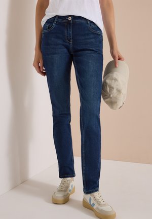 Person iført blå jeans, hvide sneakers, hvid skjorte, der holder en lysegrå baseballkasket i højre hånd.