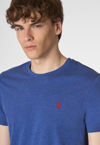 T-shirt blu a girocollo realizzato in tessuto morbido, con un piccolo logo rosso sul lato sinistro del petto. Maniche corte e vestibilità standard.