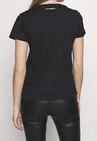 Black short-sleeve t-shirt with a round neckline, featuring a gold logo detail at the back. Worn with shiny black fitted pants.