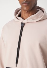 Sudadera rosa clara con un bolsillo frontal, presenta un cordón negro con un tope plateado, exhibiendo una suave textura de tela.