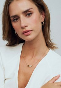 Guldhalsband med en texturerad oval berlock och en slät sfärisk accent, bärs av en person i en vit topp med v-ringad design.