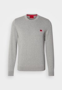 Pull gris clair à manches longues avec col rond, poignets et ourlet côtelés, arborant une petite étiquette de logo rouge sur la poitrine et une étiquette rouge à l'intérieur du col.