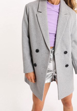Femme portant un manteau gris en laine à double boutonnage sur un crop top lavande et un short argenté métallique, debout devant un fond uni.