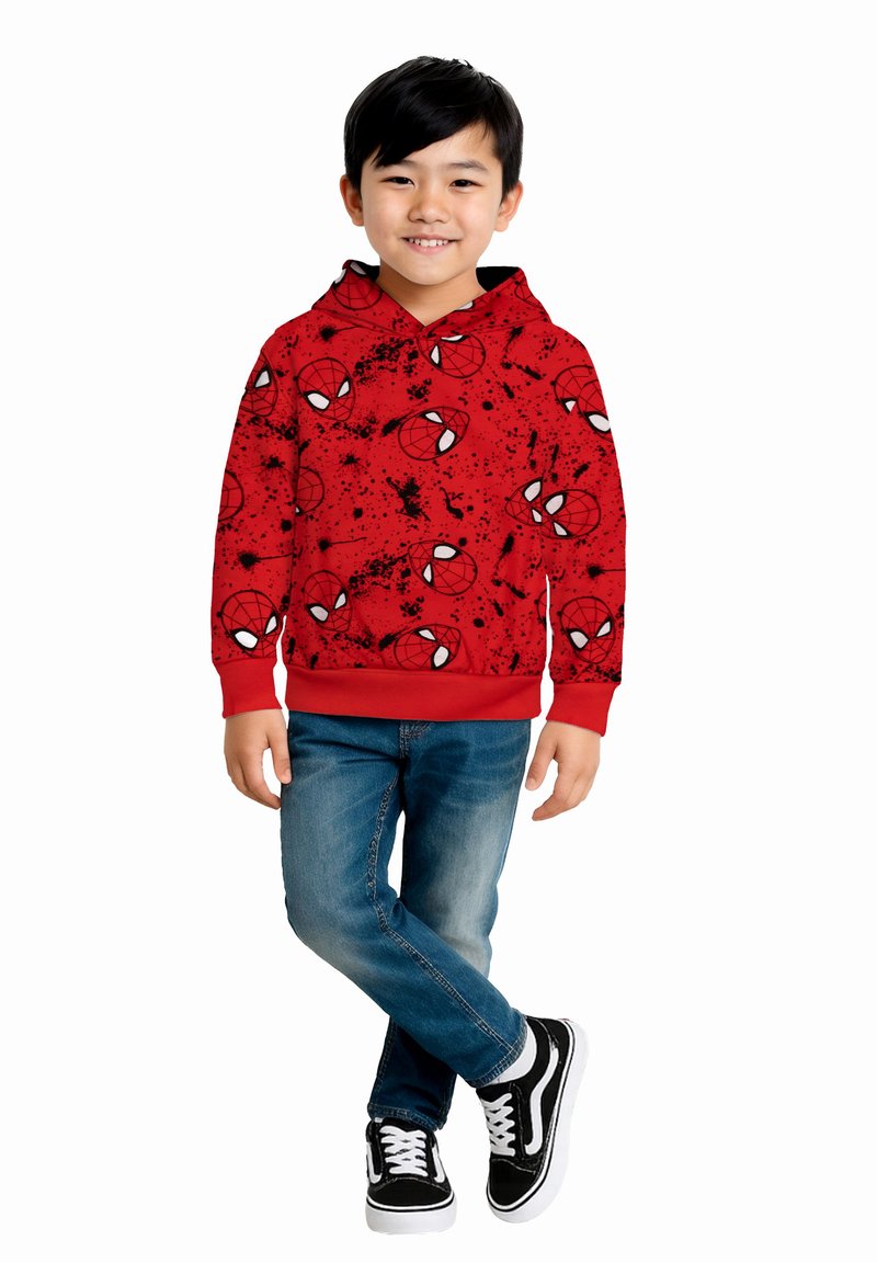 Ragazzo sorridente, che indossa un felpa rossa con disegni di ragnatela nera, jeans blu e scarpe nere, in piedi con le gambe incrociate su uno sfondo bianco.