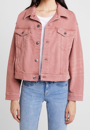 Person iført en cropped støvet lyserød denimjakke med knaplukning og klaplommer på brystet over en hvid skjorte og lyseblå jeans.