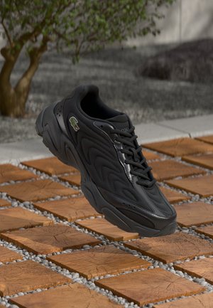 Zapatilla negra con parte superior de malla texturizada, acentos suaves y un logotipo prominente. Presenta una suela gruesa y está compuesta de materiales ligeros.