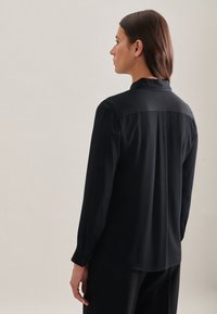 Blusa nera a maniche lunghe con una texture liscia e un design aerodinamico. Presenta un colletto classico e polsini con bottoni per la chiusura.