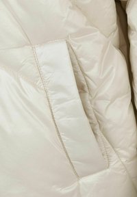 Giacca imbottita color crema con una texture liscia e lucida. Presenta un motivo a quadri diagonali e una tasca laterale, che evidenziano un design minimalista.