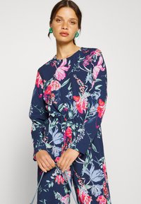 Vestido de padrão floral em tecido azul-marinho, com flores vibrantes em rosa e verde, mangas longas, decote redondo e detalhe de cintura ajustada com laço.