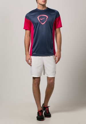 Nike Dri-FIT athletic shirt in navy and pink, short sleeves, with a rounded neck and a logo on the chest; paired with white shorts and black shoes.