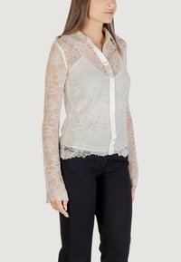 Blusa bianca in pizzo con colletto, maniche lunghe e chiusura con bottoni frontale. Presenta motivi floreali e orlo a scallop. Abbinata a pantaloni neri.