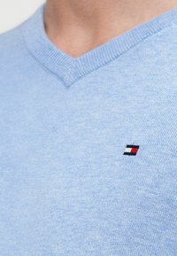 Ανοιχτό μπλε πουλόβερ με λαιμόκοψη σε σχήμα V από μαλακό πλεκτό ύφασμα, με μικρό κεντημένο λογότυπο σε κόκκινα, λευκά και μπλε στοιχεία στο στήθος.