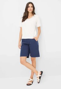 Witte kortekraagtop met kantafwerking, gecombineerd met knielange marineblauwe shorts. Geddraagd met lichtgekleurde sandaaltjes met bandjes. Eenvoudig ontwerp, casual stijl.