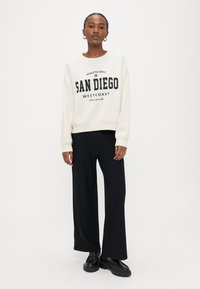 Frau trägt weißen Pullover mit dem Schriftzug "San Diego West Coast", schwarze weite Hosen und schwarze Loafers, steht vor weißem Hintergrund.