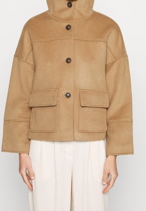 Veste en laine beige avec boutons sur le devant, grandes poches à rabat, manches longues et col haut portée sur un pantalon blanc.