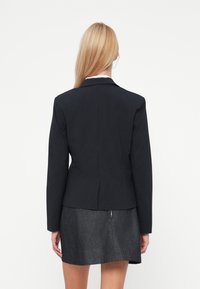 Blazer noir à rayures avec une coupe ajustée, un col et des épaulettes ; associé à une jupe en denim foncé, mettant en valeur un zip à l'arrière.