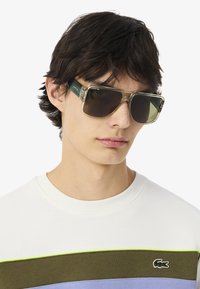 Gafas de sol con montura transparente y acentos verdes, con lentes oscuros rectangulares. Llevadas por un modelo que viste un sudadera multicolor.