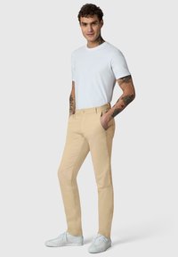 Jeune homme aux cheveux bouclés portant un T-shirt blanc, un pantalon beige, des baskets blanches et affichant des tatouages sur les bras et le cou, debout les mains dans les poches.