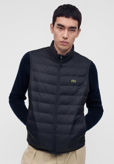 Homme portant un gilet matelassé noir avec un petit logo crocodile vert, des manches longues noires en dessous et un pantalon beige, sur un fond uni.