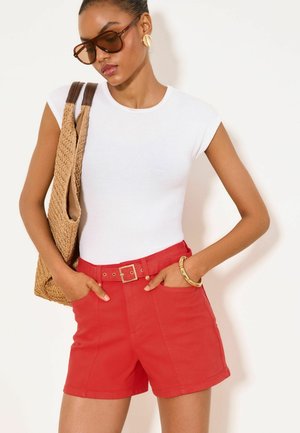 Kobieta nosząca brązowe okulary przeciwsłoneczne, biały top bez rękawów, czerwone spodenki z paskiem, złotą bransoletkę i kolczyki, nosząca na ramieniu beżową plecioną torbę.