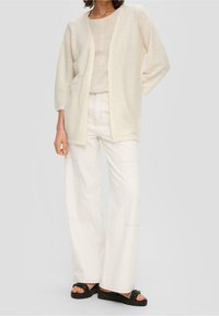 Světle béžový kardigan s otevřeným předním dílem, volným střihem a tříčtvrtečními rukávy, doplněný béžovým topem a širokými bílými kalhotami. Černé sandály.