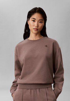 Ung kvinde med glat mørkt hår bærer en brun Calvin Klein sweatshirt og matchende bukser, står mod en ensartet lys grå baggrund.