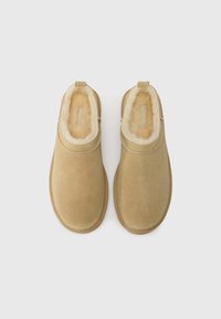 Scarpe slip-on in suede beige con una morbida fodera in pelliccia color crema, punta arrotondata e dettagli di cucitura sottile lungo il bordo superiore.