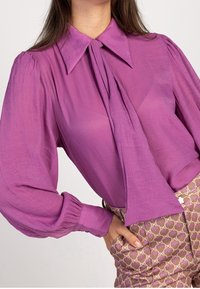 Blusa lunga viola con colletto ampio, dettaglio a cravatta al collo e tessuto testurizzato; abbinata a pantaloni a vita alta con fantasia.