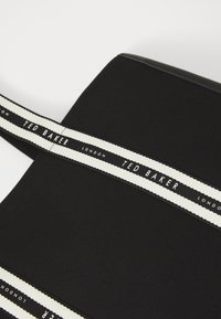 Schwarze Stofftasche mit strukturiertem Oberflächend esign, akzentuiert mit einem weißen Trageriemen, der im wiederholten Muster "TED BAKER LONDON" beschriftet ist.