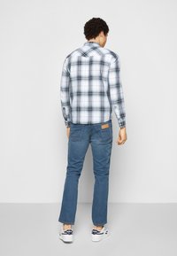 Camicia da uomo a quadri blu e bianca, con polsini abbottonati, abbinata a jeans blu. Caratteristiche notevoli includono una spalla posteriore e un'etichetta in pelle color cognac.