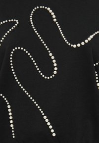 Top en tissu noir orné d'accents blanc en forme de perles disposés en lignes ondulées, créant un motif décoratif texturé.