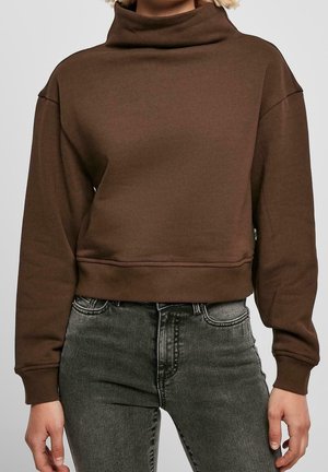 Personne portant un sweat-shirt court marron à col roulé et manches longues et un jean noir délavé taille haute, debout les bras détendus.