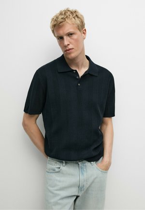 Junger Mann mit kurzen blonden Haaren, der ein schwarzes, geripptes kurzärmeliges Poloshirt und hellblaue Jeans trägt, steht vor einem weißen Hintergrund.