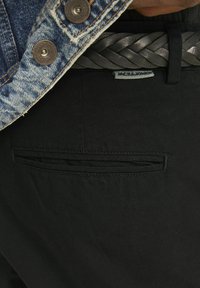 Sorte stofbukser med en glat tekstur, en baglomme og et flettet sort læderbælte med en sølvspænde. En denimjakke synlig ovenpå.