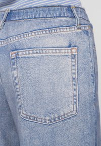 Svetlomodré deninové džínsy so zadnou vreckom, s kontrastným oranžovým prešívaním a mierne vyblednutou textúrou. Viditeľný detail pásu.