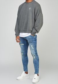 Sudadera gris de gran tamaño con un logotipo bordado en blanco, combinada con jeans ajustados de color azul claro desgastados y zapatillas blancas.