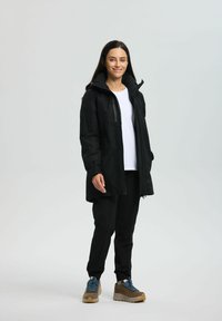 Schwarze wasserdichte Jacke mit Kapuze, Frontreißverschluss und zwei Seitentaschen, getragen über einem weißen Hemd und schwarzen Hosen, kombiniert mit braunen Wanderschuhen.