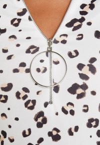 Vitt tyg med bruna och svarta leopardfläckar. Har en silverfärgad dragkedja med ett runt draghandtag vid halsringningen. Slät textur.